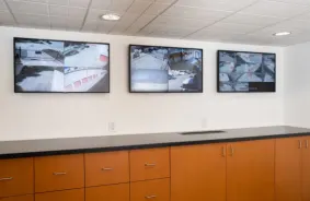 Three TV screens mounted on the wall display surveillance footage of the facility.
