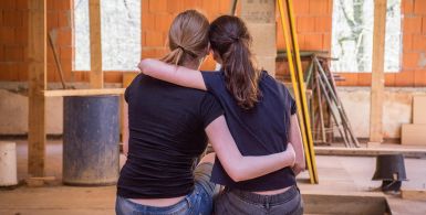 back side view of two women hugging looking at a house being remodeled from the inside