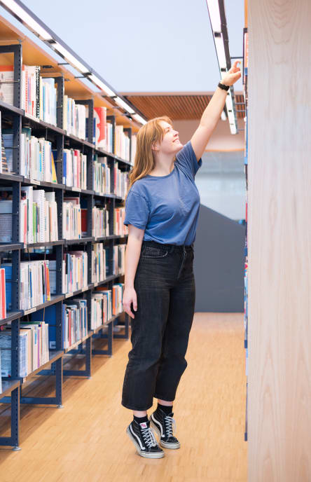 Student gjør tåhev ved bokhyllene på biblioteket USN Campus Vestfold