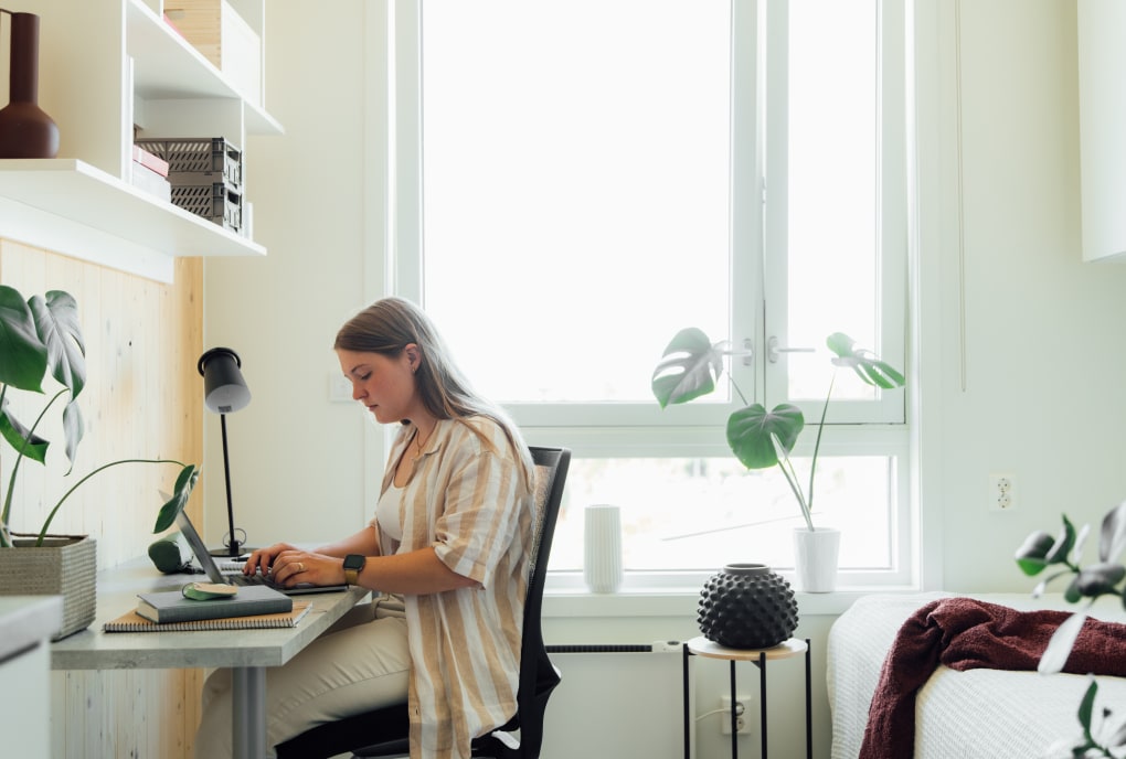 Student sitter ved pult på hybel og skriver på en laptop