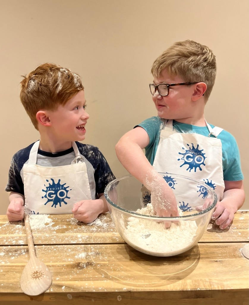 Two young brothers, Owen and George. mixing flour wearing Project GO aprons