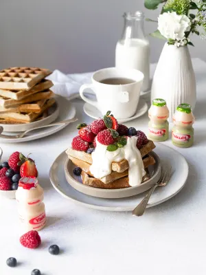 Vanilleboekweitwafels met vers fruit, mascarponesaus, kopje thee, melk, bloemen in een vaasje met flesjes Yakult Plus en Original.