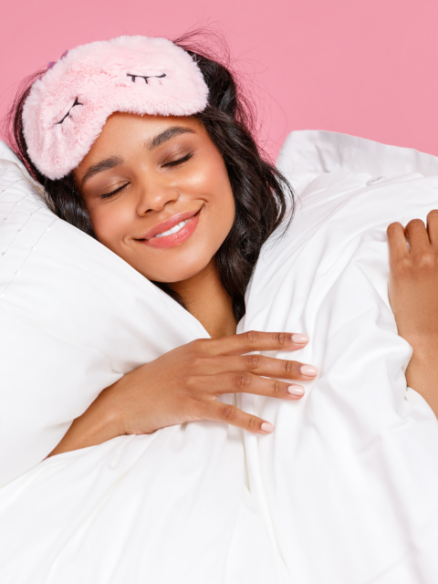 A woman smiling, wearing a pink, fluffy eye mask and holding pillows against a pink background