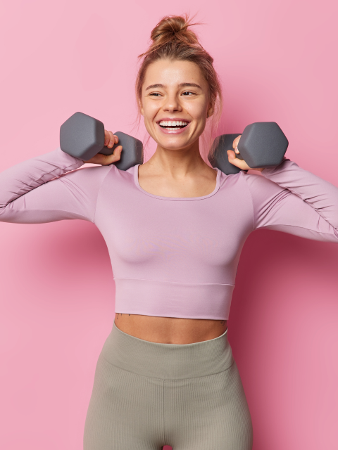 A woman standing, smiling against a pink background as she lifts dumbbells above her shoulders