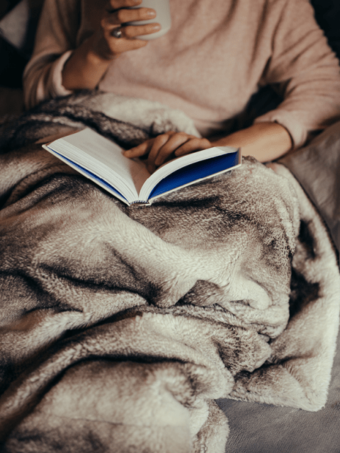 Frau mit Buch und Teetasse, eingekuschelt in Decke