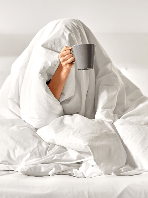 Frau, die sich unter der Bettdecke versteckt und eine Kaffeetasse in der Hand hält.