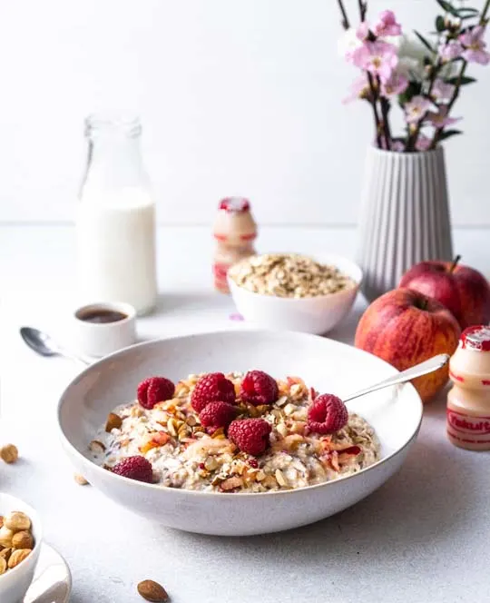 Muesli met frambozen in een kom, fles melk, kopje koffie,appels, noten, bloesem in een vaasje en een flesje Yakult Original.