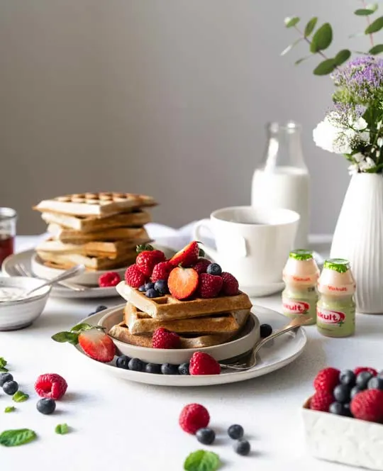 Vanilleboekweitwafels met vers fruit, mascarponesaus, kopje thee, melk, bloemen in een vaasje met flesjes Yakult Plus.