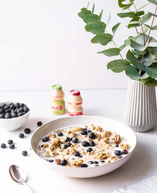 Granola met blauwe bessen en flesjes Yakult Original, Plus met een kommetje blauwe bessen.