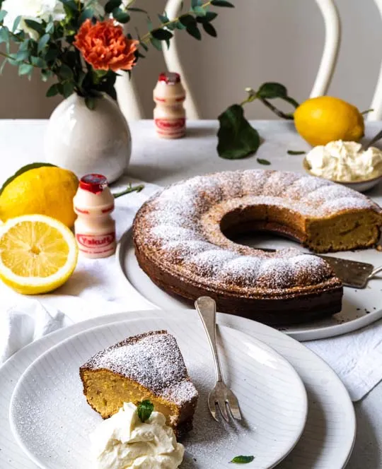 Citroen ontbijtcake met een flesje Yakult Original op een tafel met een boeket en citroenen.