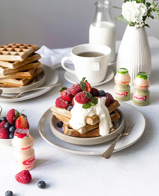 Vanilleboekweitwafels met vers fruit, mascarponesaus, kopje thee, melk, bloemen in een vaasje met flesjes Yakult Plus en Original.