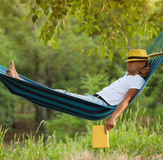 Man sleeping in striped hammock wearing a yellow hat and holding a yellow book