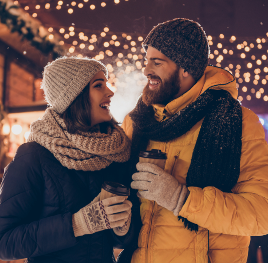 A happy couple smiling at each other, holding a coffee cup in theirs hands and wearing woolly hats and scarves