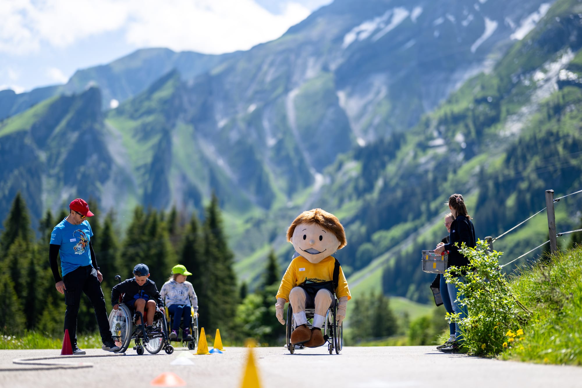 Kinder im Rollstuhl mit Maskottchen im Rollstuhl vor Bergpanorama