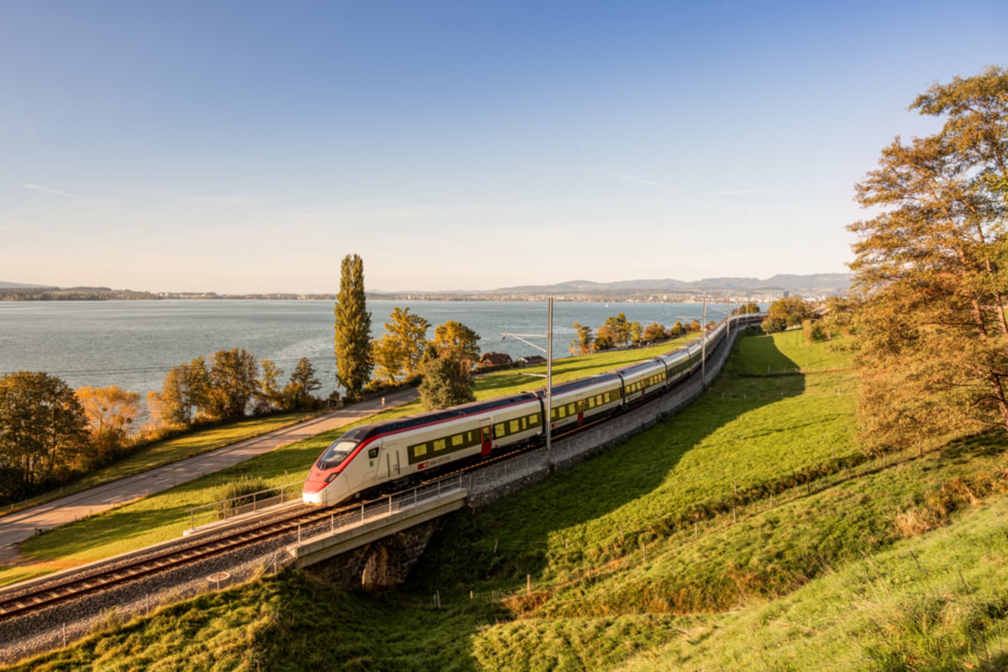 Zug fährt über eine herbsliche Wiese, im Hintergrund ein See