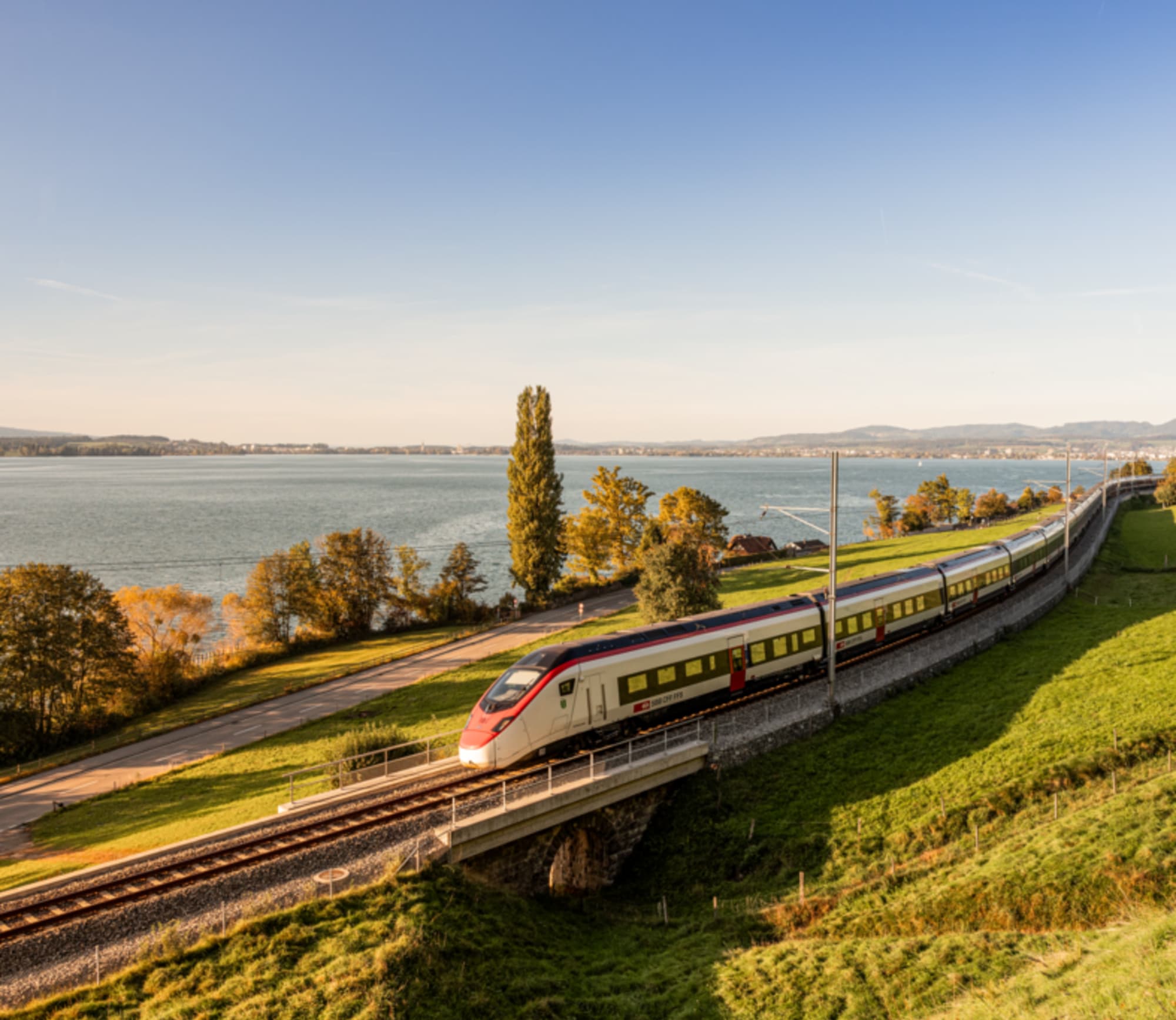 Zug fährt über eine herbsliche Wiese, im Hintergrund ein See