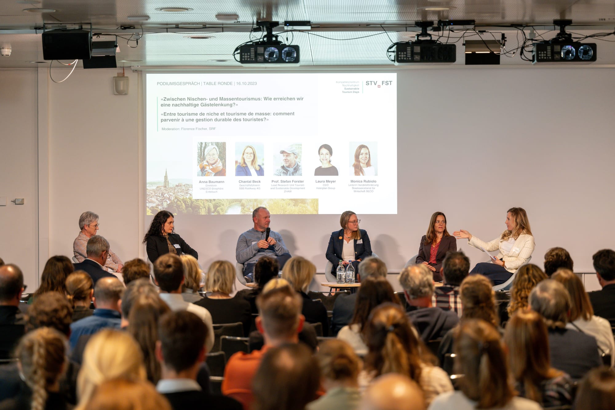 Foto eines Podiumsgesprächs, auf der Bühne sitzen sechs Personen, im Vordergrund des Fotos sieht man das Publikum von hinten, Foto von den Sustainable Tourism Days 2023