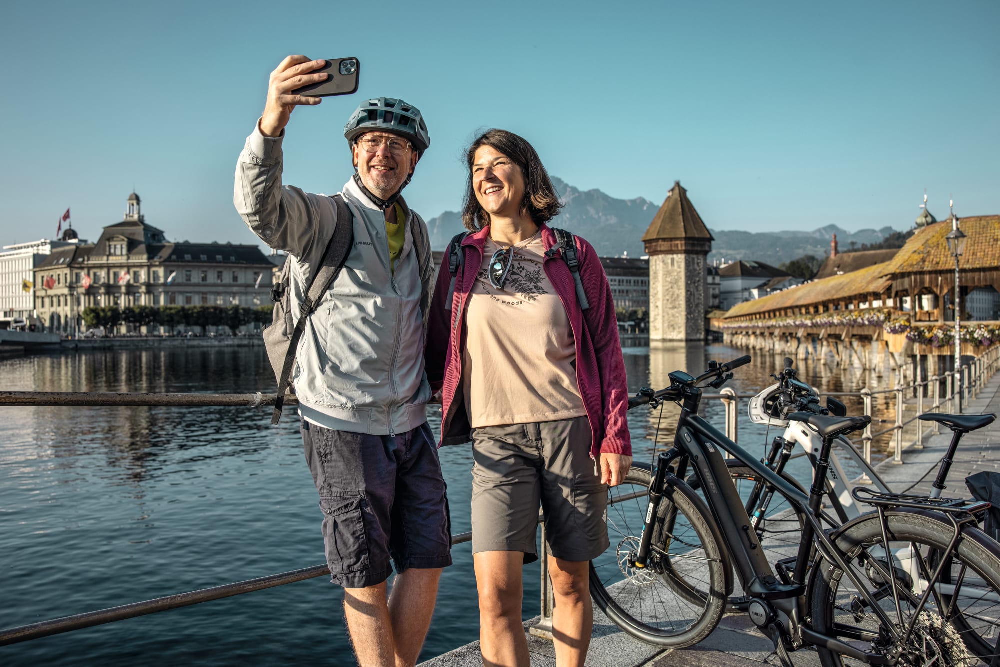 Un homme, une femme et des vélos prennent un selfie devant le pont Kappelbrücke et le mont Pilate.