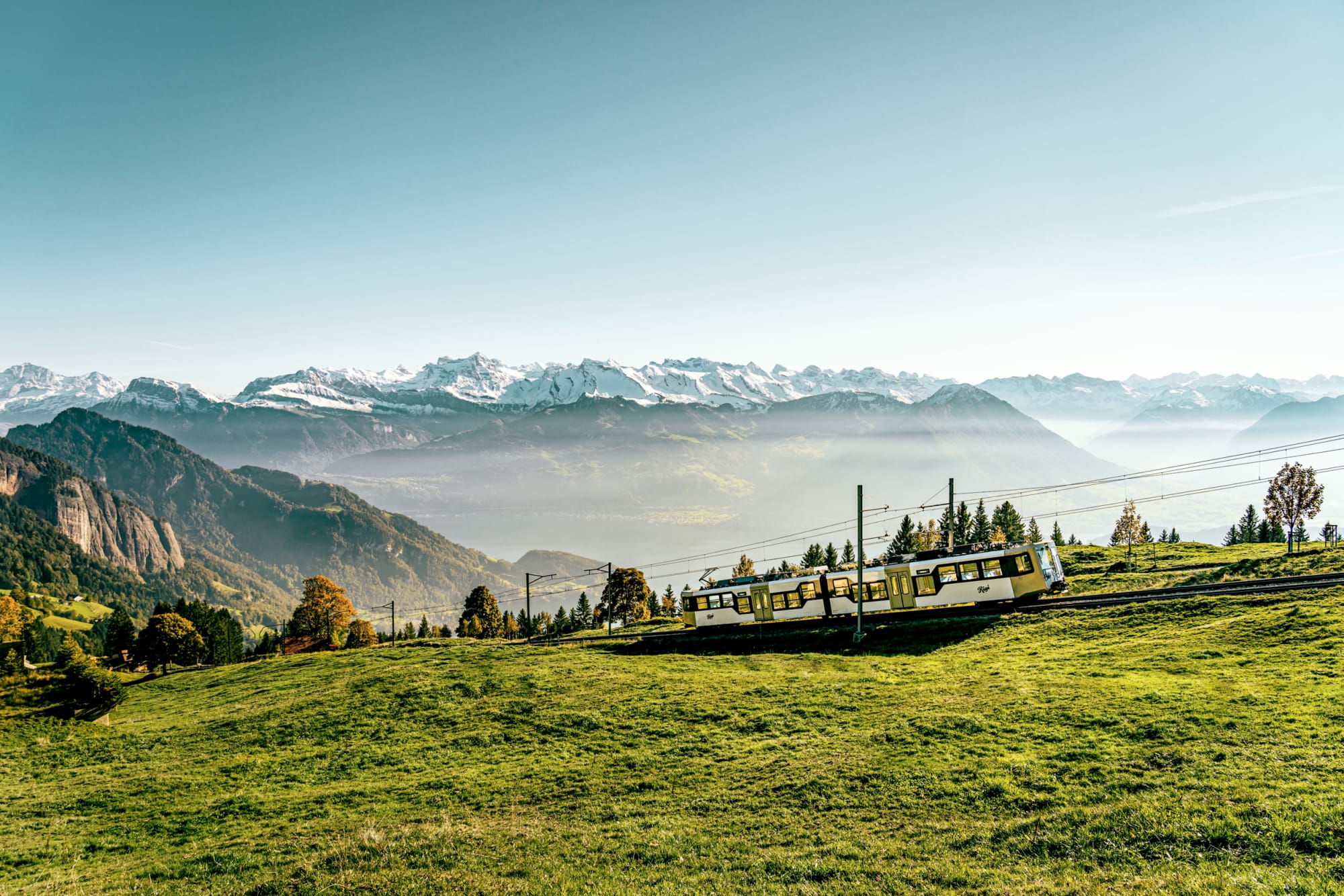 Die neue Rigi Bahn fährt den Berg hoch. Im Hintergrund sind die Berge zu sehen bei schönem Wetter und etwas Nebel.