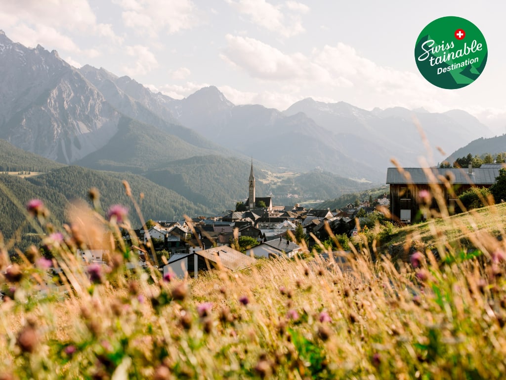 Blick auf das Dorf Sent im Unterengadin mit einem Bergpanorama im Hintergrund, Swisstainable Destination Logo in der oberen rechten Bildhälfte.