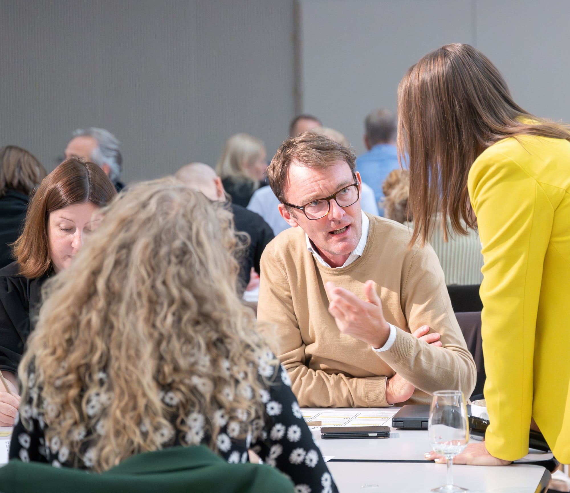 Personen tauschen sich am Workshop aus: mehrere Personen sitzen, eine steht auf den Tisch gestützt da