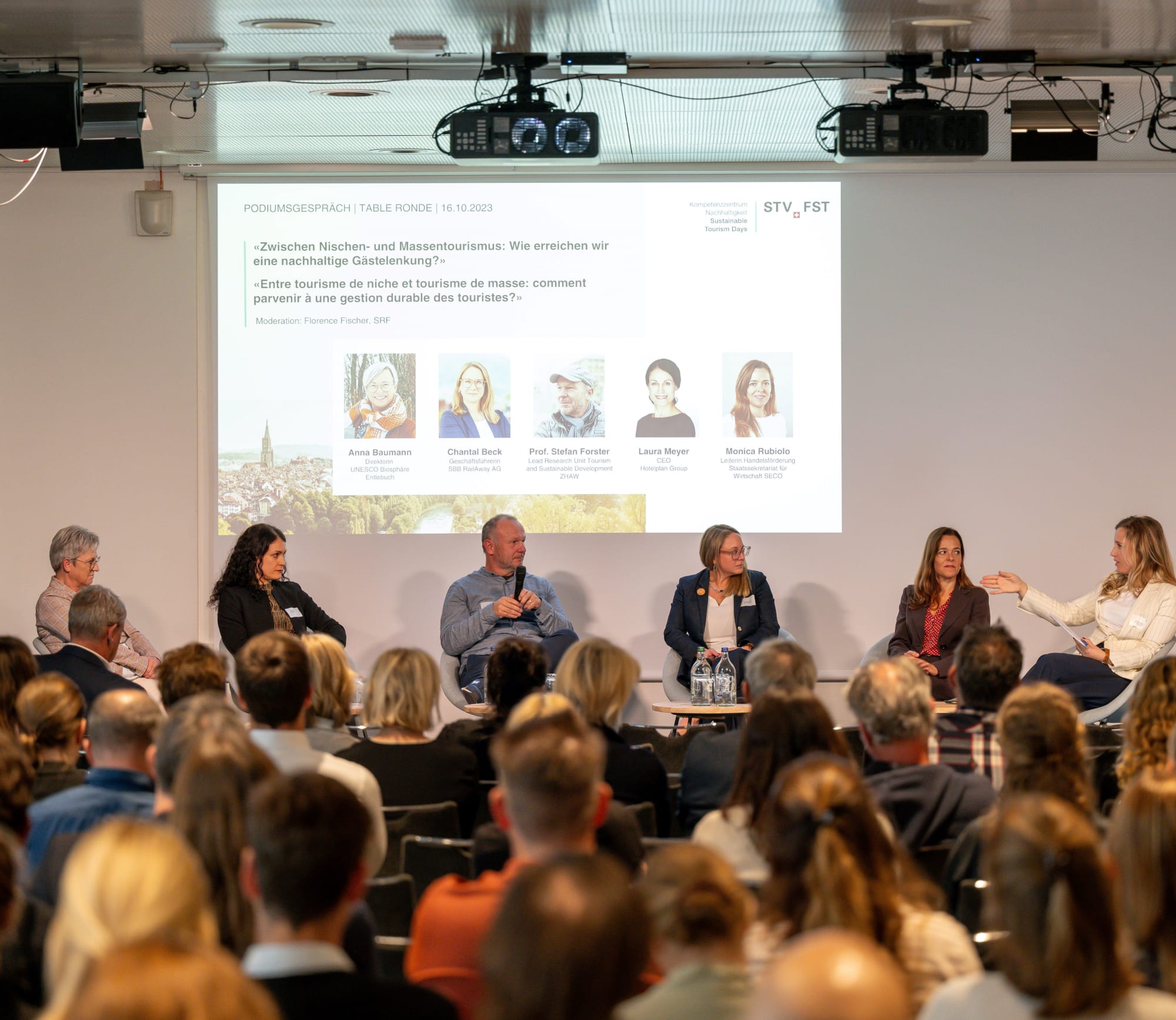 Foto eines Podiumsgesprächs, auf der Bühne sitzen sechs Personen, im Vordergrund des Fotos sieht man das Publikum von hinten, Foto von den Sustainable Tourism Days 2023