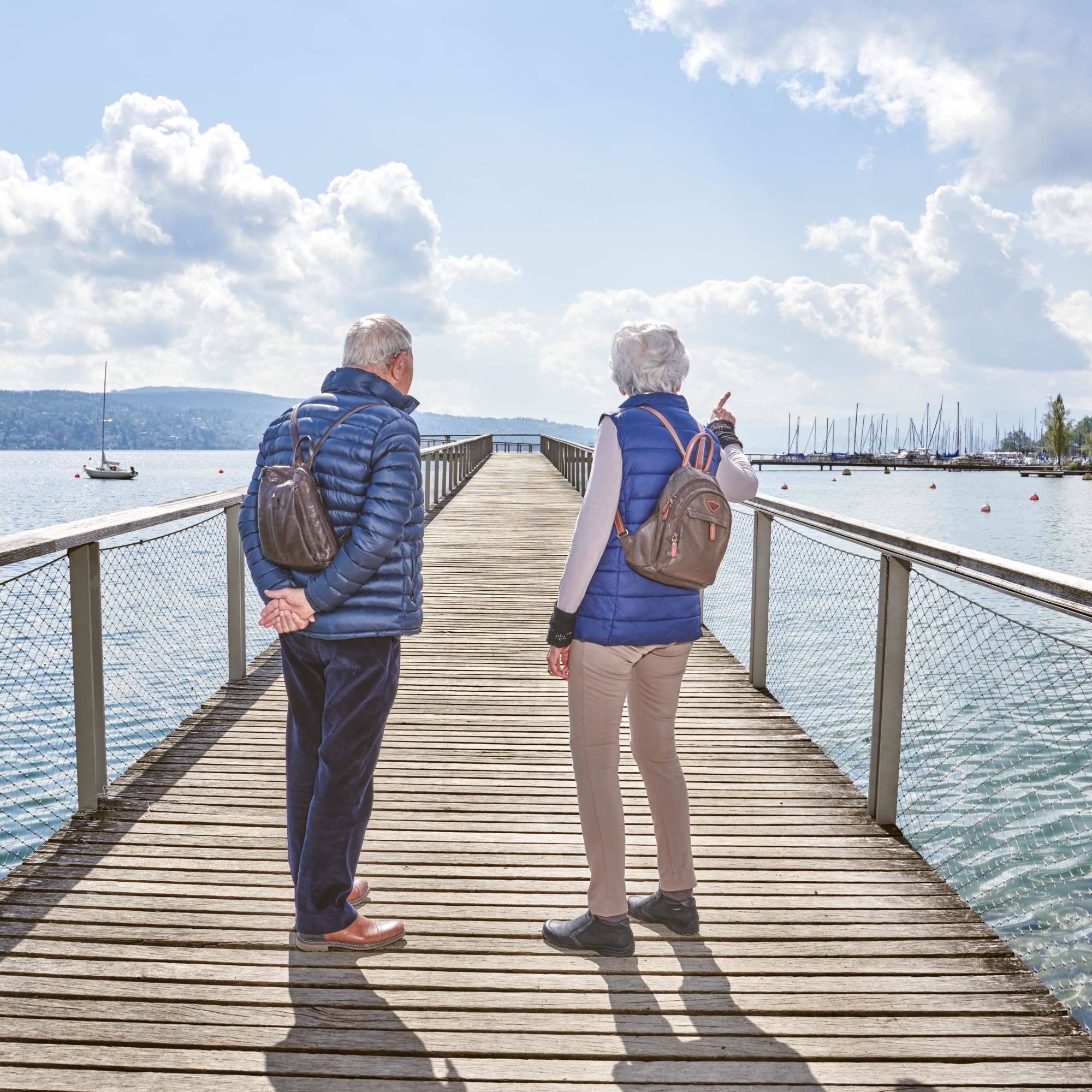 Zwei Senioren stehen auf einer Brücke, vor ihnen ein See mit Segelbooten. Die Frau zeigt in die Ferne
