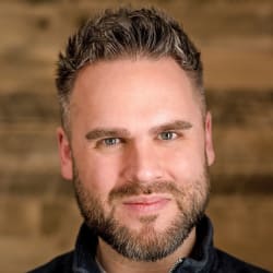 Headshot of Dean Breyley, bearded, in front of a wooden wall