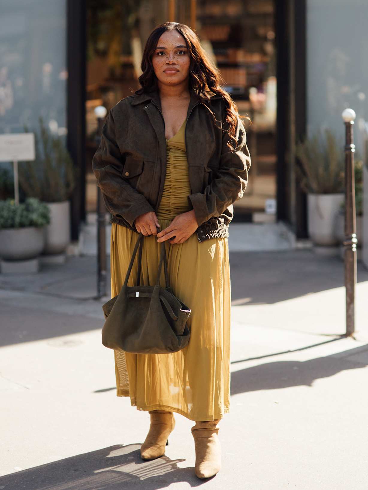 Frau steht vor einem GEschäft und trägt eine dunkle Lederjacke und ein gelbes Kleid, in der Hand hält sie den Shopper THE NEW YORK LARGE von DEMELLIER aus grünem Suede.