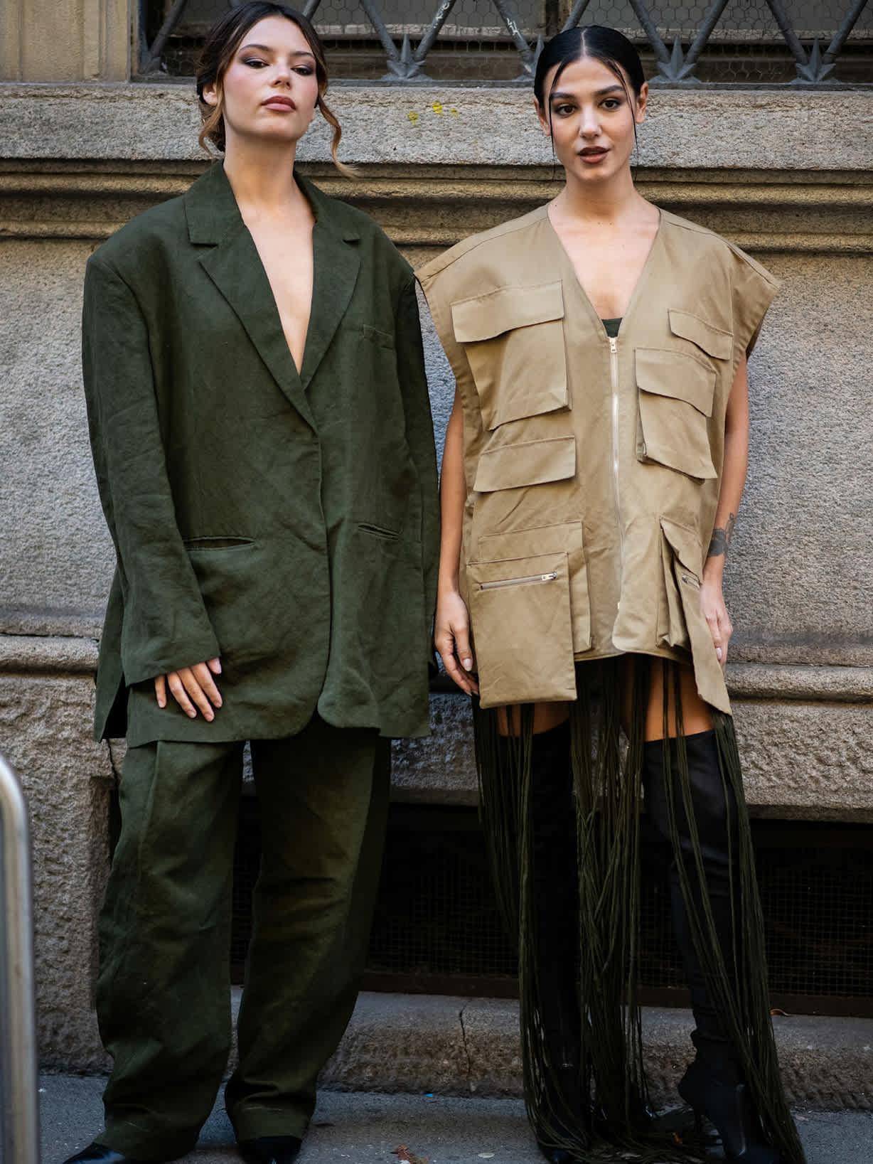 Zwei Frauen stehen vor einer Mauer, eine Frau trägt einen grünen Blazer zu einer grünen Hose und die andere eine beige Weste mit aufgesetzten Taschen  und Overknees