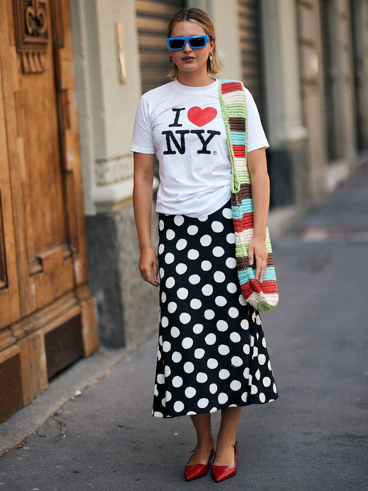Frau trägt einen schwarz-weiß gepunkteten Rock, dazu ein weißes T-Shirt mit der Aufschrift I love NY, dazu rote Ballerinas und eine blaue Sonnenbrille
