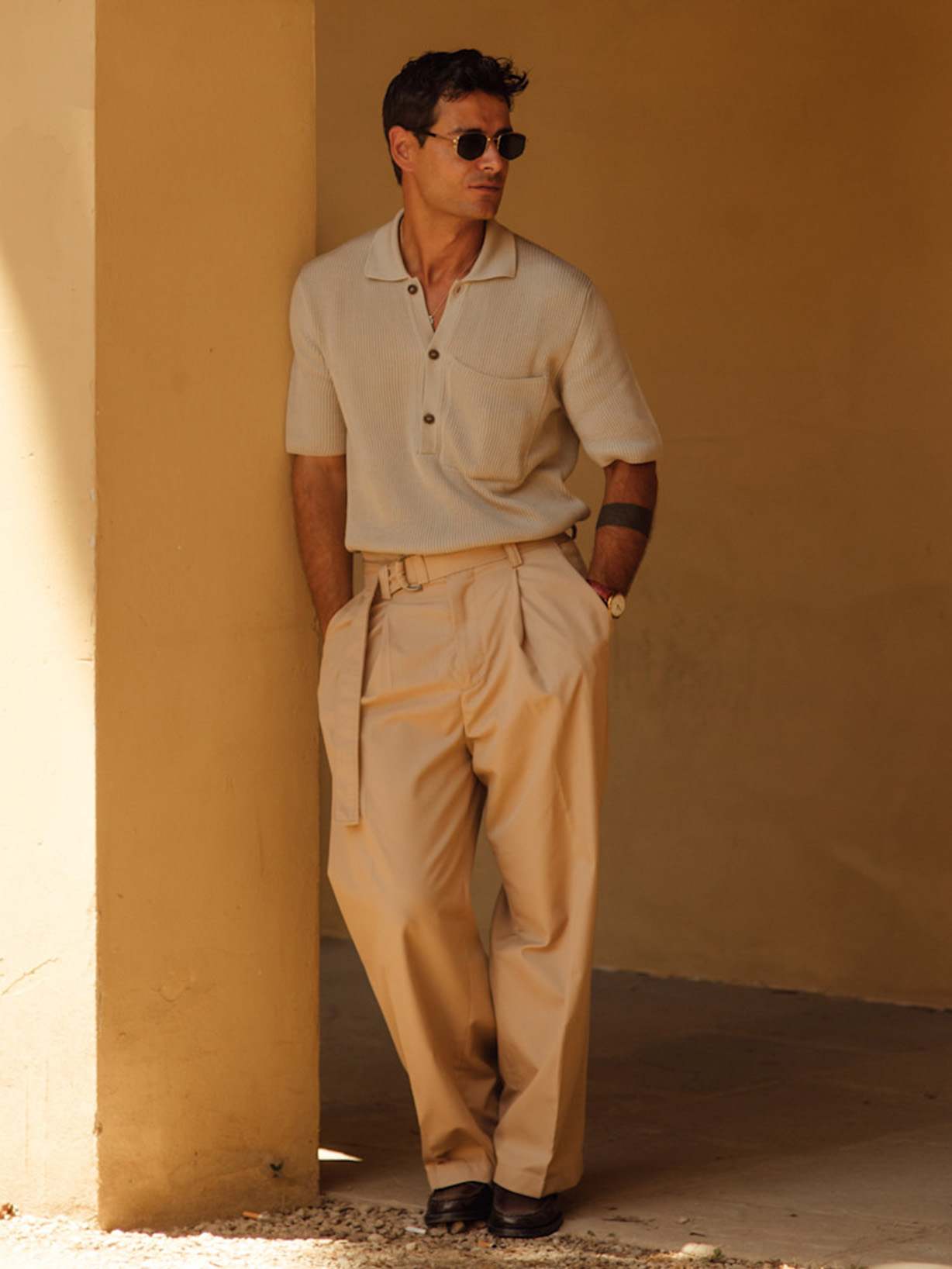 Mann stylte beige Bundfaltenhose und hellem Kurzarm-Strickpolo, dunklen Loafern und Sonnenbrille