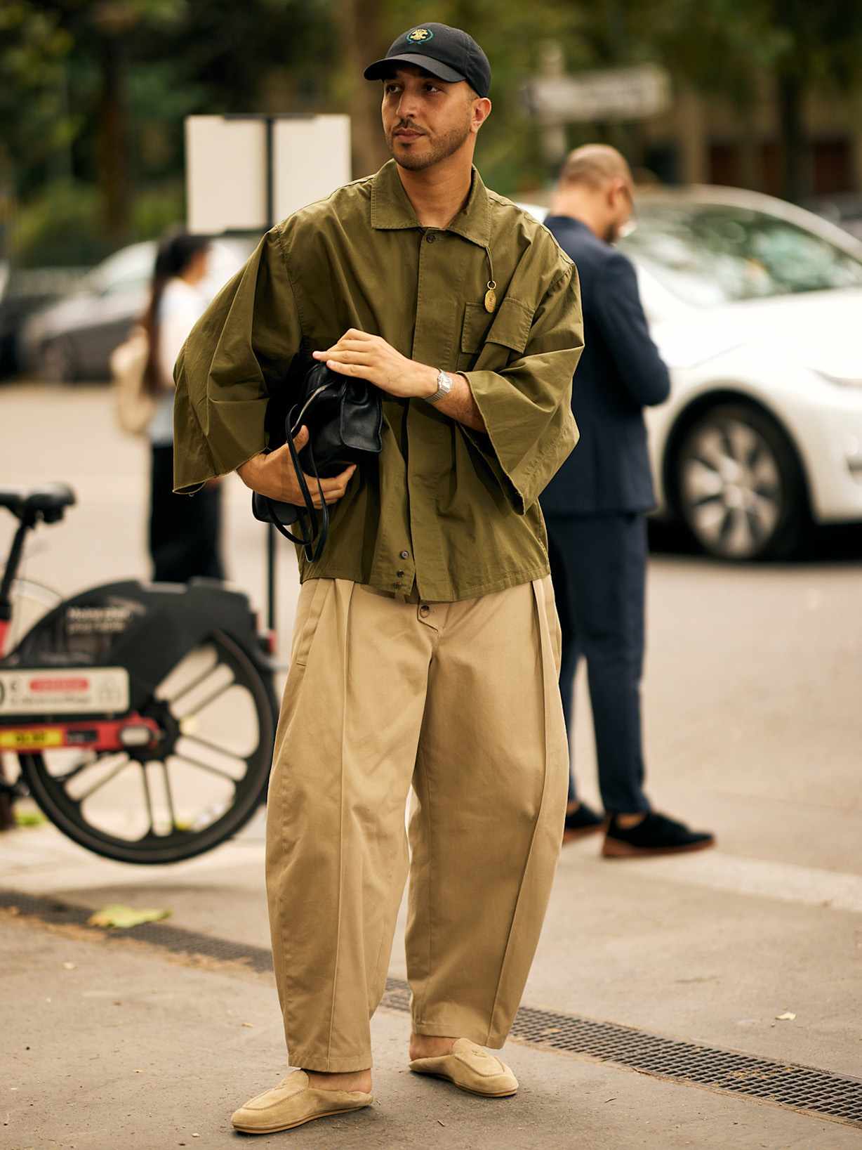 Mann in weiter beiger Hose und olivgrünem Overshirt, kombiniert mit Cap, heller Slip on Silhouette und schwarzer Tasche