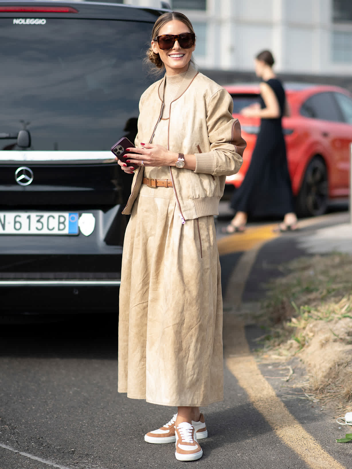 Frau in beigefarbenem Bomberjacken-Look mit langem A-Linien-Rock, Sonnenbrille und Sneakern