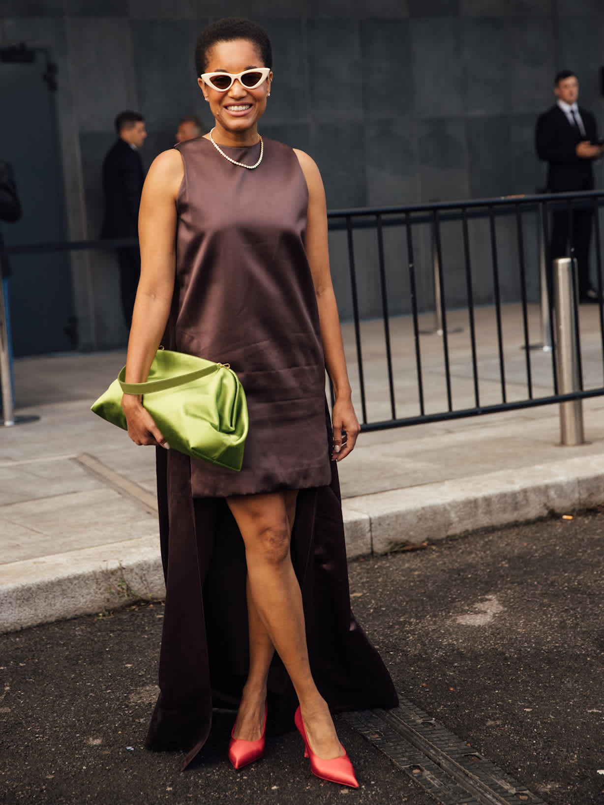 Frau in dunkelbraunem Satinkleid mit langer Rückseite, grüner Clutch, weißer Sonnenbrille und roten Pumps