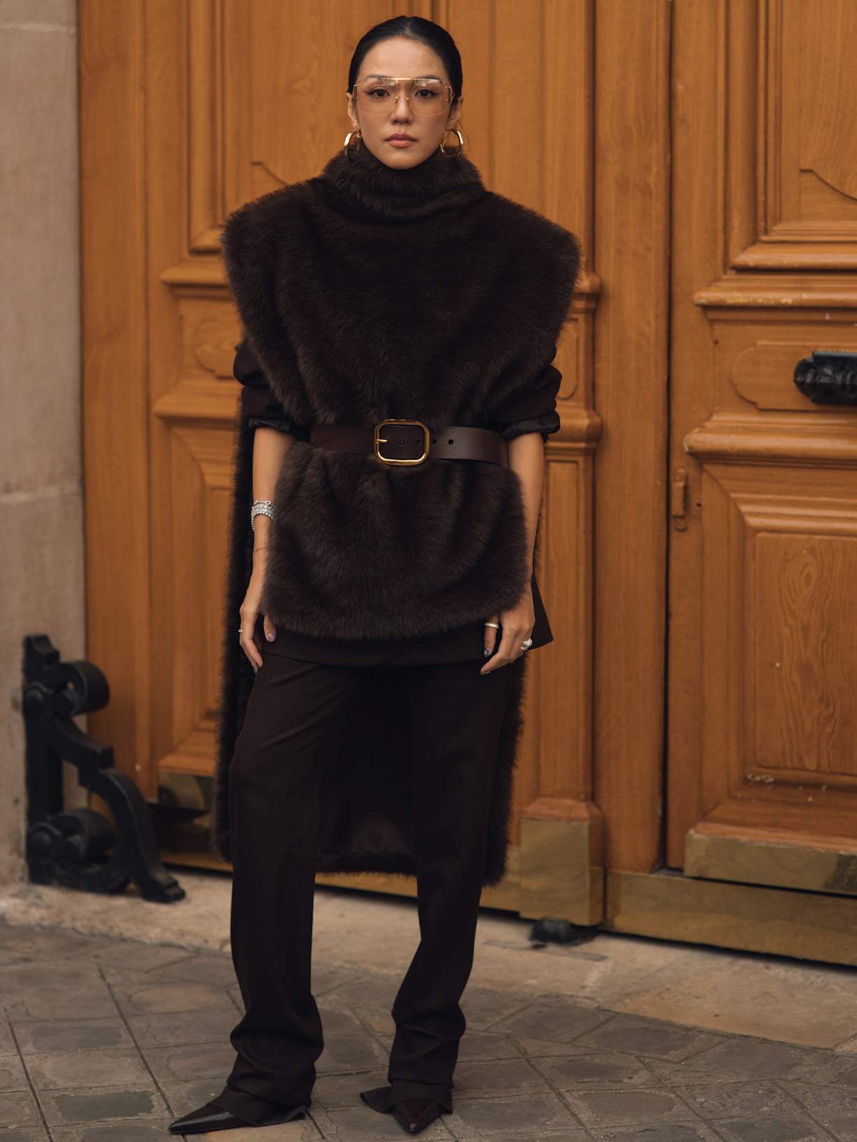 Eine Frau trägt ein dunkelbraunes Outfit mit ärmellosem Kunstfell-Weste, breitem Taillengürtel, schmaler Stoffhose und spitzen Schuhen.