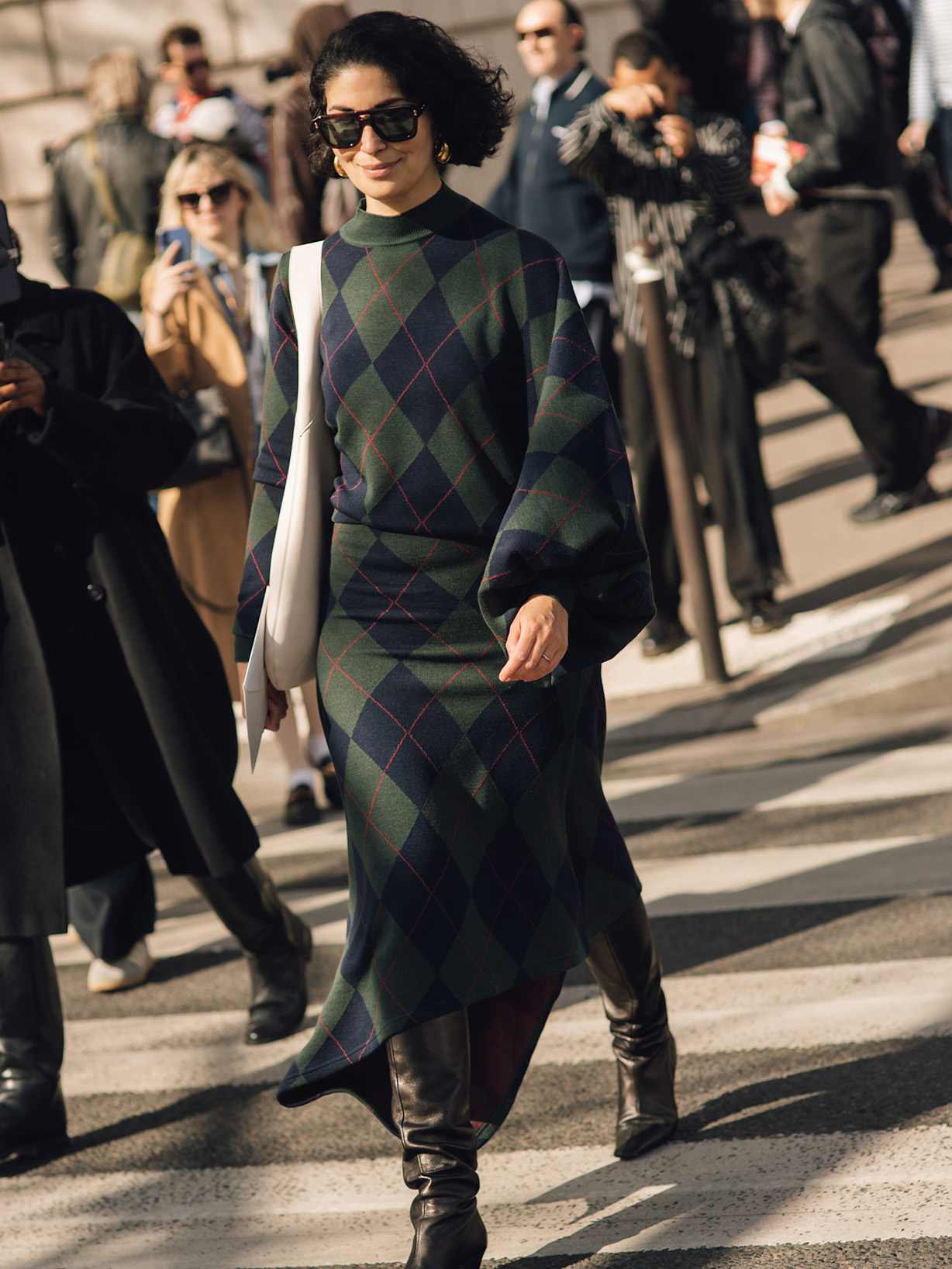 Eine dunkelhaarige Frau trägt Herbst-Outfit mit dunkelgrünem Strickkleid im Rautenmuster, kniehohen schwarzen Lederstiefeln und weißer Schultertasche