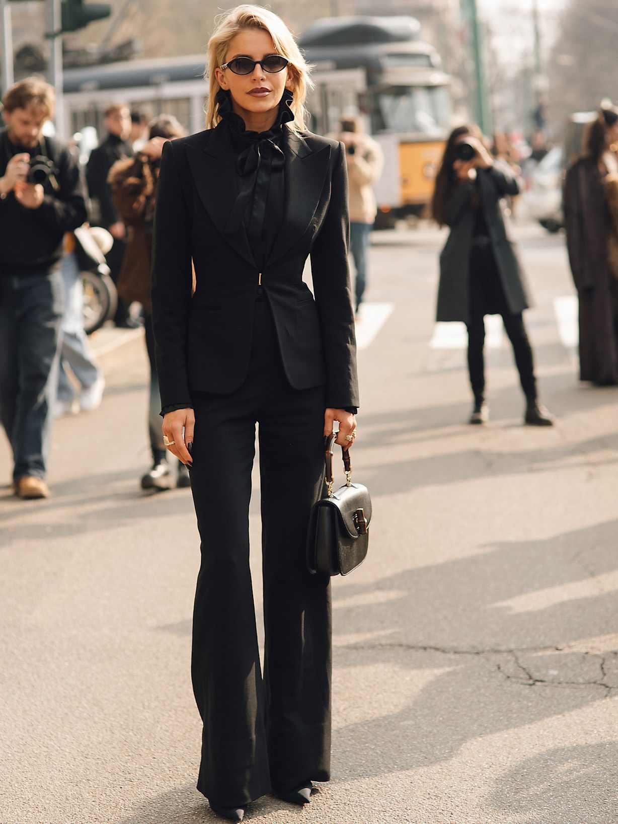 Caro Daur in einem eleganten schwarzen Hosenanzug mit tailliertem Blazer, Schluppenbluse, Sonnenbrille und schwarzer Handtasche auf einer belebten Straße.