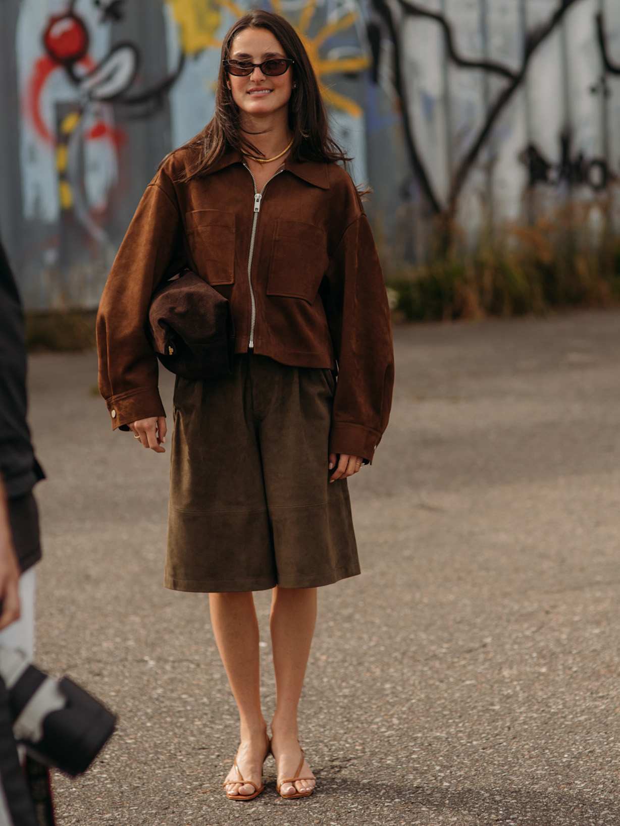 Frau in brauner Wildlederjacke, khakifarbenen Bermudashorts und Sonnenbrille