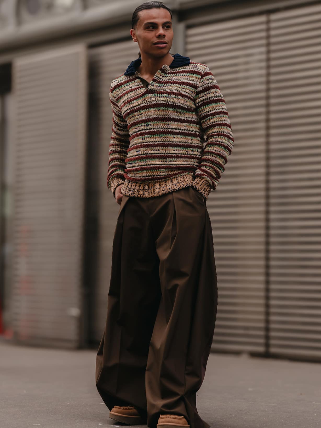Mann mit Grobstrickpullover mit einer weiten braunen Hose steht auf der Straße. 