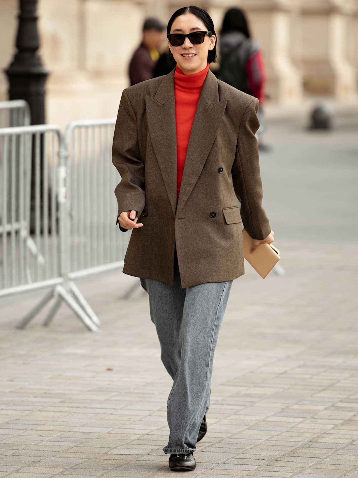 Frau trägt einen roten Strickpullover unter einem braunen Blazer und zu einer grauen Stoffhose. 