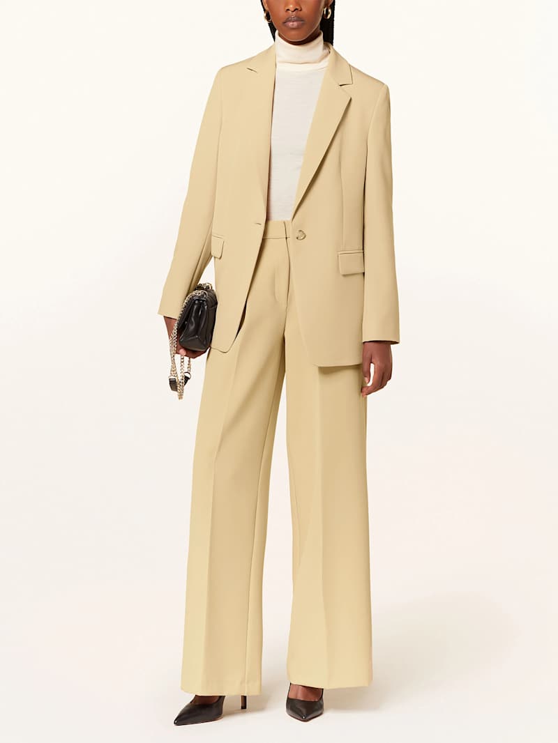 Frau trägt eine beige Business-Kombination mit nudefarbenem Rollkragenpullover und schwarzer Clutch