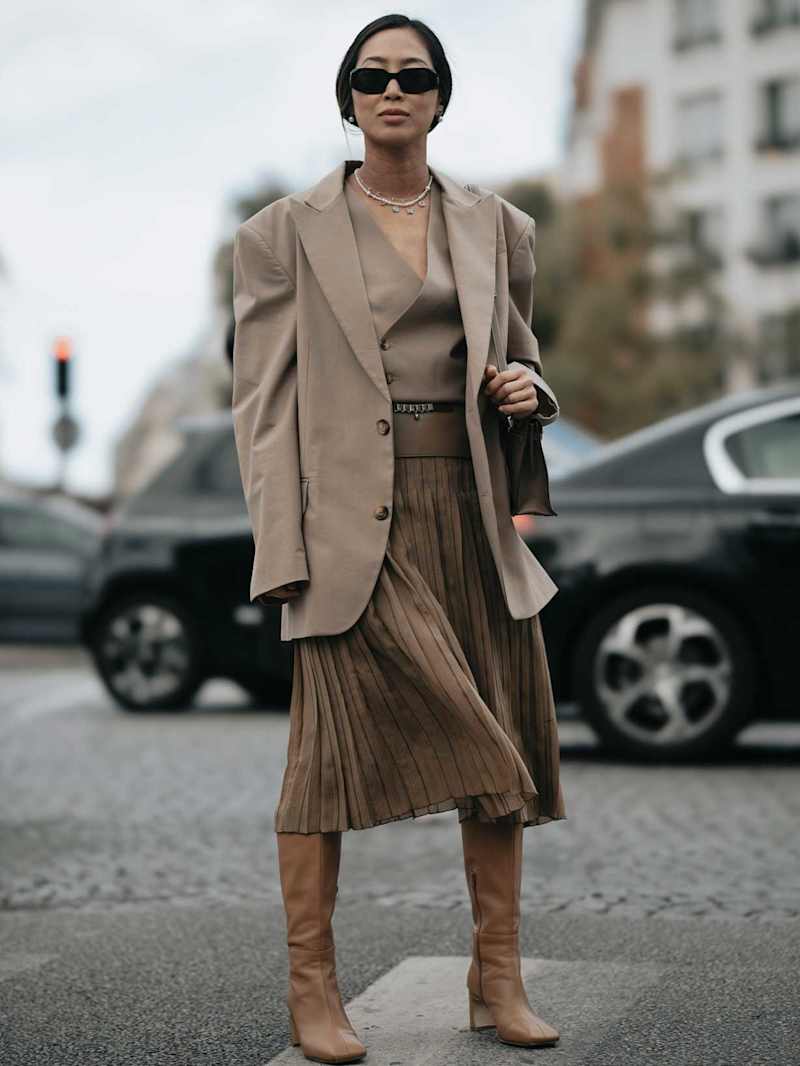 Frau trägt ein monochromes Outfit in beige bestehend aus Top, Blazer, Plisseerock und kombiniert dazu Stiefel