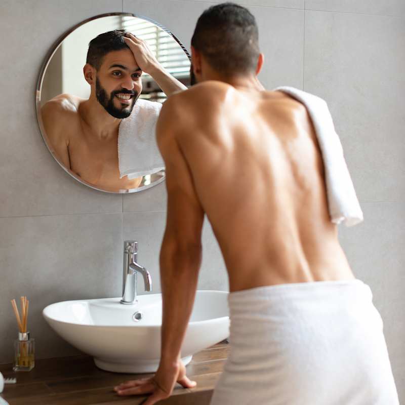 Dunkelhaariger Mann mit Bart betrachtet seine Haare im Spiegel