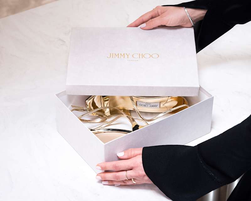 شخص يفتح صندوق حذاء أبيض من Jimmy Choo يحتوي على صندل ذهبي في بوتيك الفاخر Breuninger ميونخ