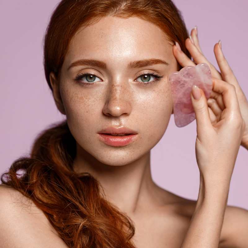 Rothaarige Frau behandelt ihr Gesicht mit einem Gua Sha Stein aus Rosenquarz