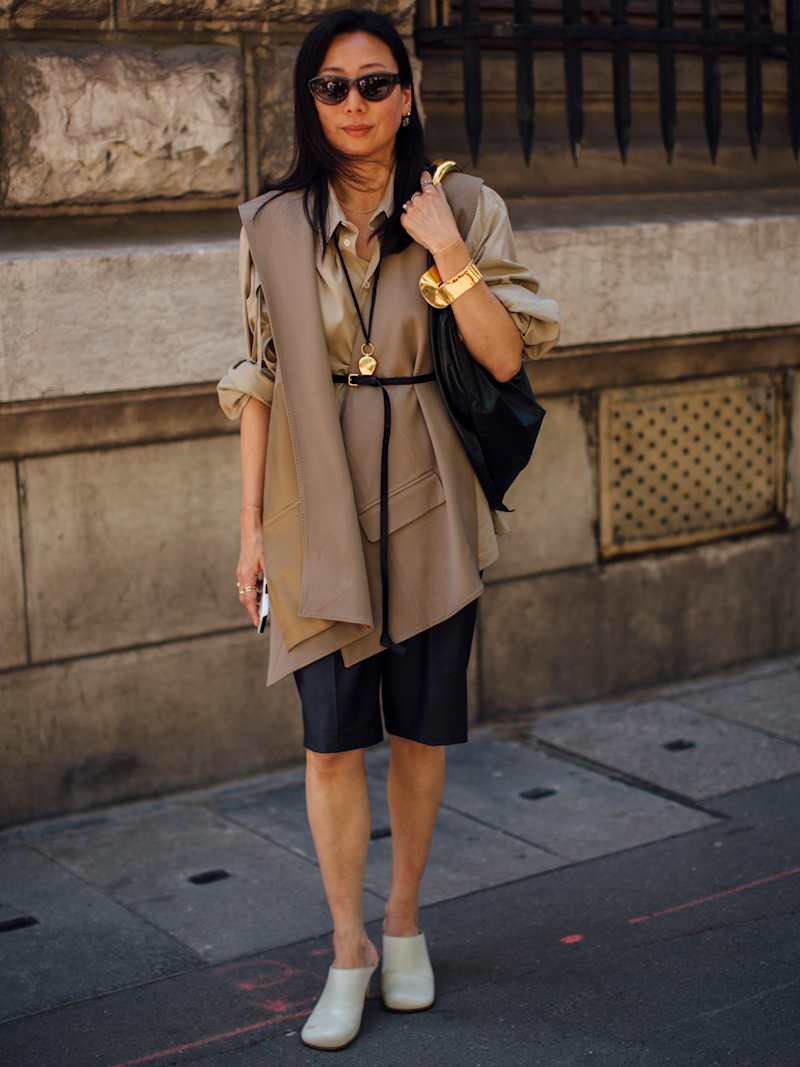 Frau trägt eine beige Weste über einer beigen Bluse zu schwarzen Bermudas und Mules in Creme, dazu goldene Accessoires