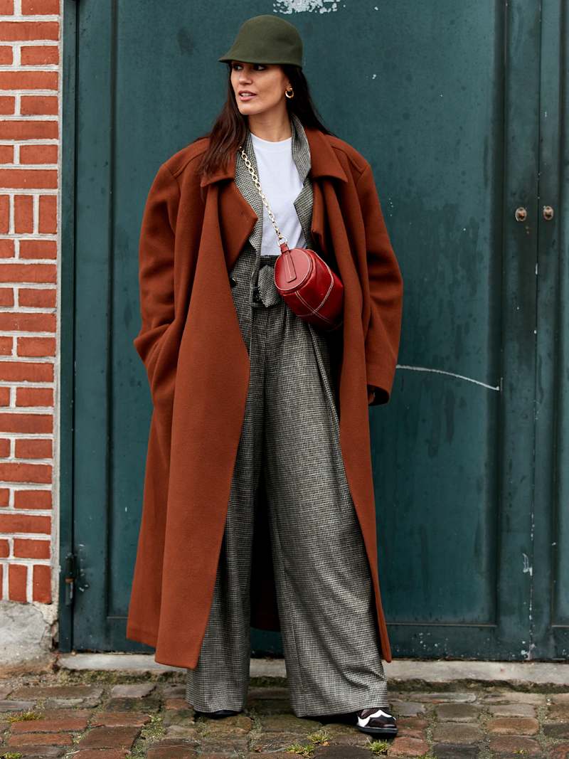 Frau trägt einen roten Wollmantel über einer Kombination aus Blazer und Hose aus grauem Tweed-Stoff, dazu einen grüne Wollfilz-Kappe