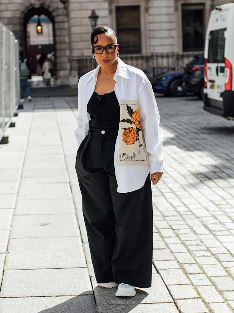Frau trägt ein schwarzes Top zu einer schwarzen, weit geschnittenen Stoffhose und darüber eine weiße Bluse mit einem großen Blumen-Print