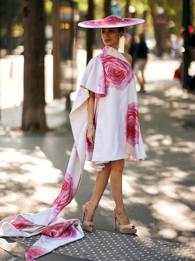 Frau trägt ein weißes Cocktailkleid mit Rosen als großflächiges Blumenmuster, dazu High Heels und einen eleganten Hut